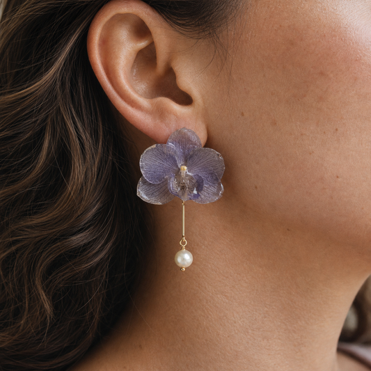 A close-up of a woman's ear wearing a dangling earring. The earring features a translucent lavender-purple orchid flower with a small clear faceted bead at the center and a dangling white pearl on a gold-toned hook.