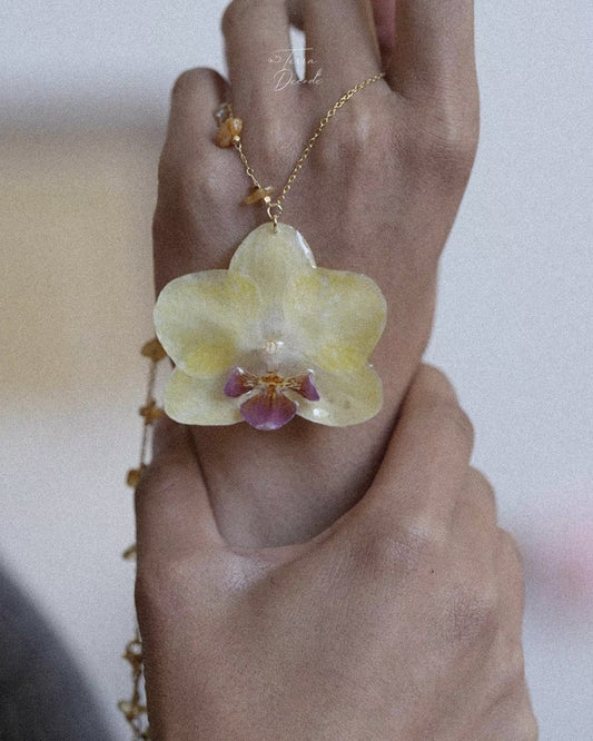 A close-up of a hand wearing a delicate gold chain necklace with a translucent pale yellow orchid flower pendant and small gold beads along the chain.