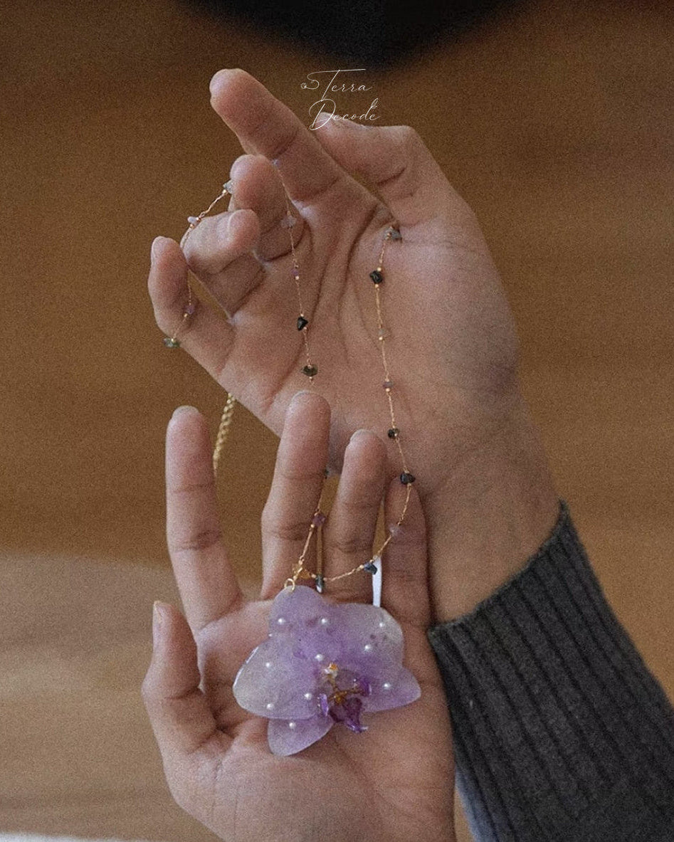 A person's hands holding a delicate gold chain necklace with a translucent purple orchid flower pendant, set against a warm wooden background.