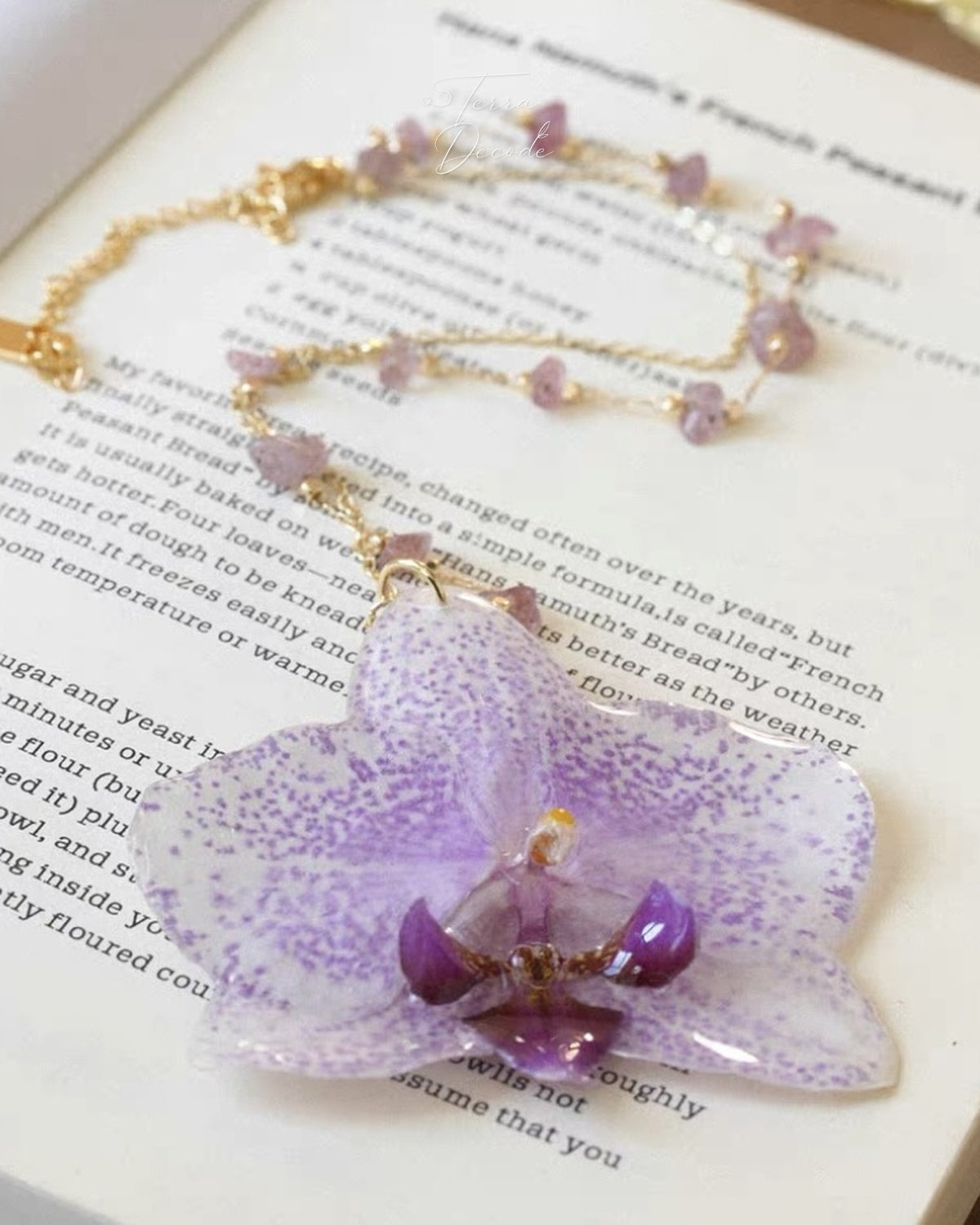 A delicate gold chain necklace with small pinkish-purple beads and a large translucent purple orchid flower pendant, displayed on an open book.
