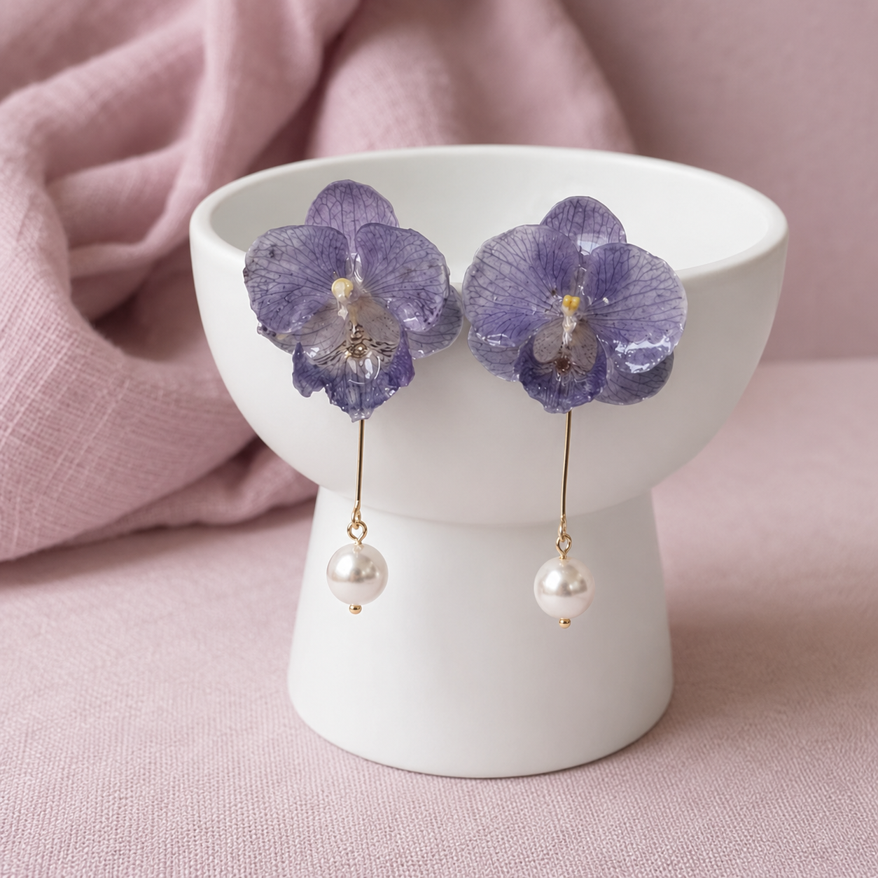 Purple orchid flower drop earrings with pearl-like dangling beads and gold-tone hooks, displayed on a white pedestal against a soft pink fabric background.