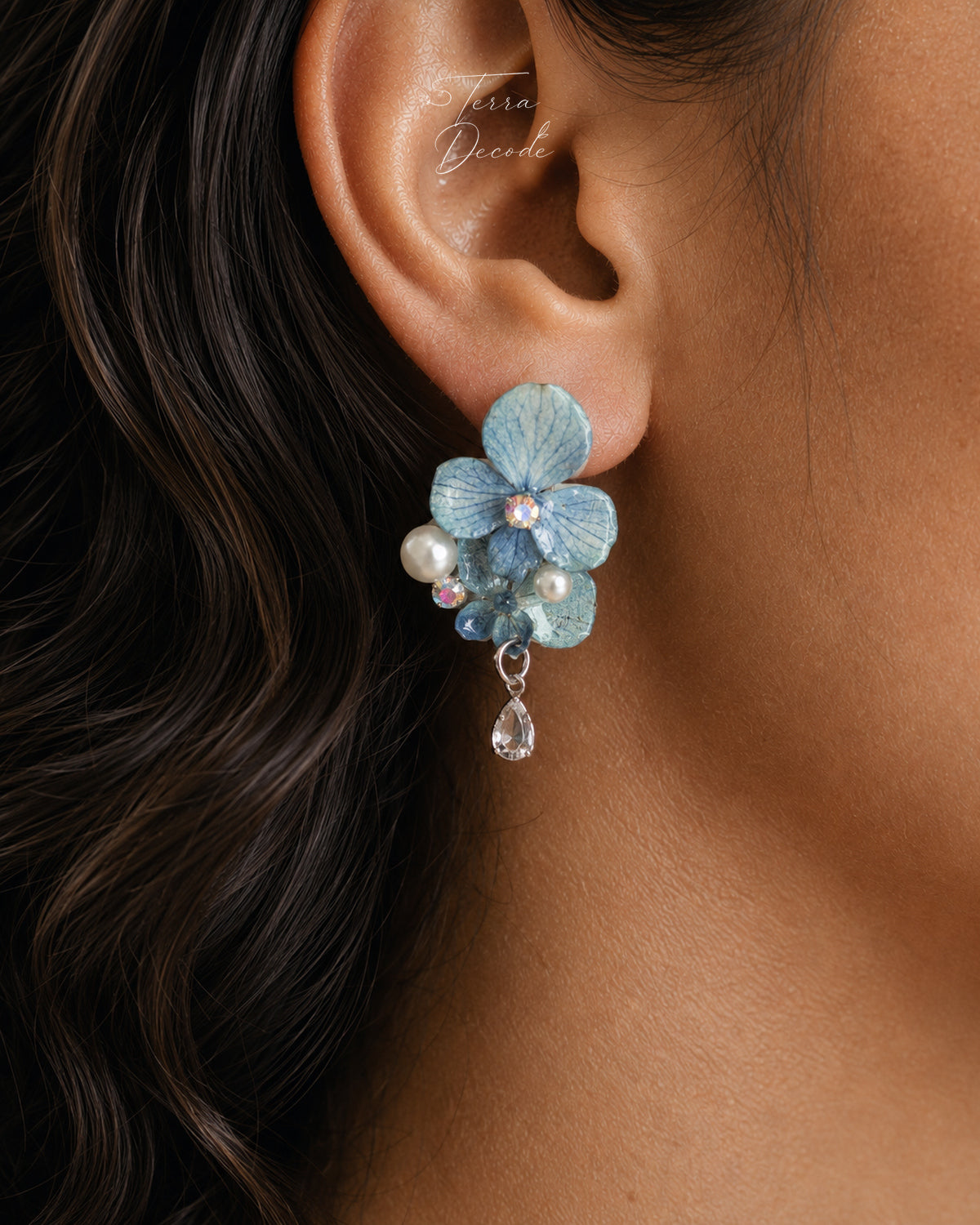 A close-up of a model's ear wearing a light blue flower-shaped hydrangea dangle earring with pearl-like beads and a clear teardrop crystal, against dark brown hair and warm skin tones.