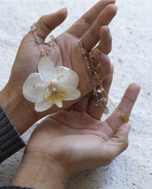 A delicate gold chain necklace featuring a large white orchid flower pendant with a yellow center and small iridescent beads, held in two hands against a textured light gray background.