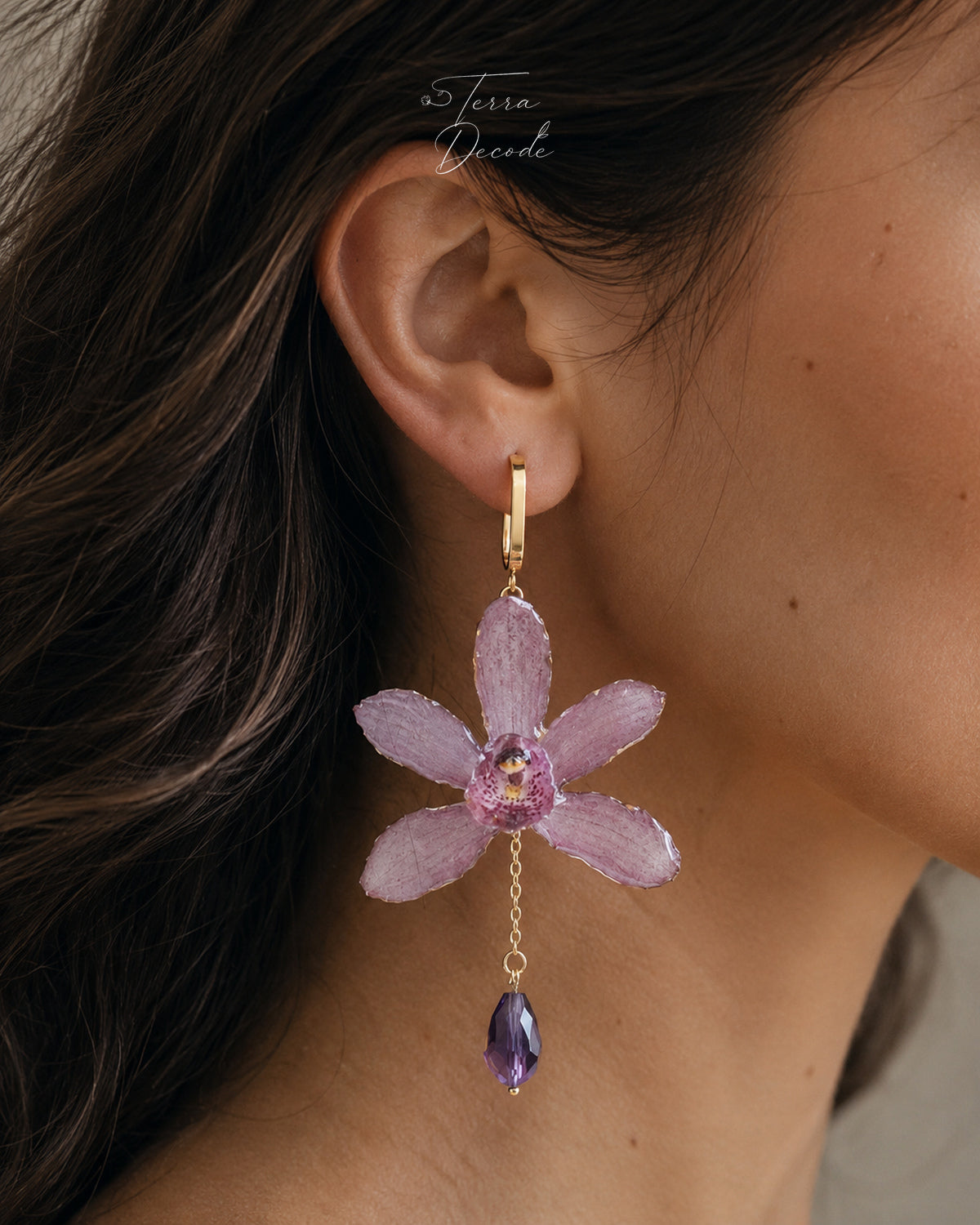 A close-up of a model's ear wearing a dangling flower earring. The earring features a gold hoop and a lavender-pink orchid flower with a purple center and a dangling purple teardrop gemstone.